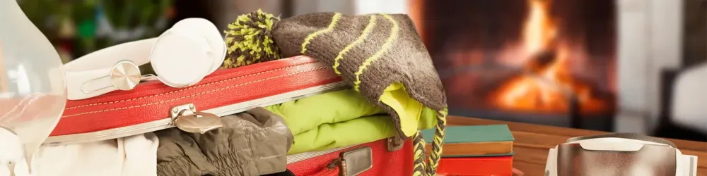 Close up of suitcase full of clothes, with a stocking hat and headphones on top, sitting in front of a fireplace